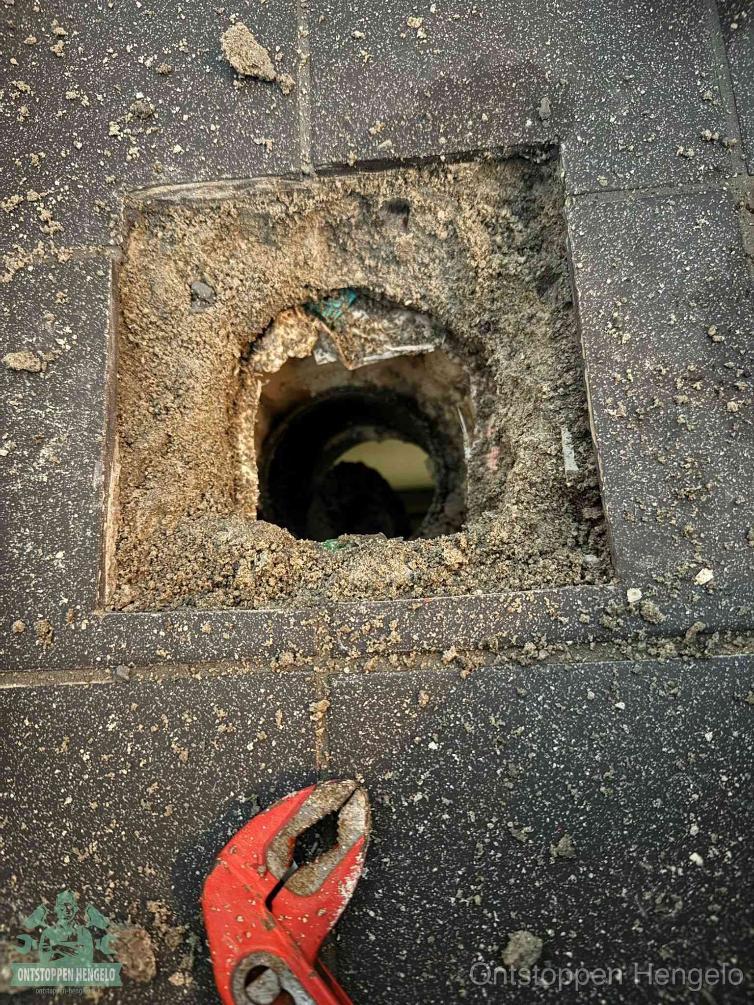 Een geopend rioolputje in een betontegelvloer met losgemaakt rooster en een zichtbare ronde afvoerbuis, het achterhalen waar de afvoerverstopping zit in Hengelo