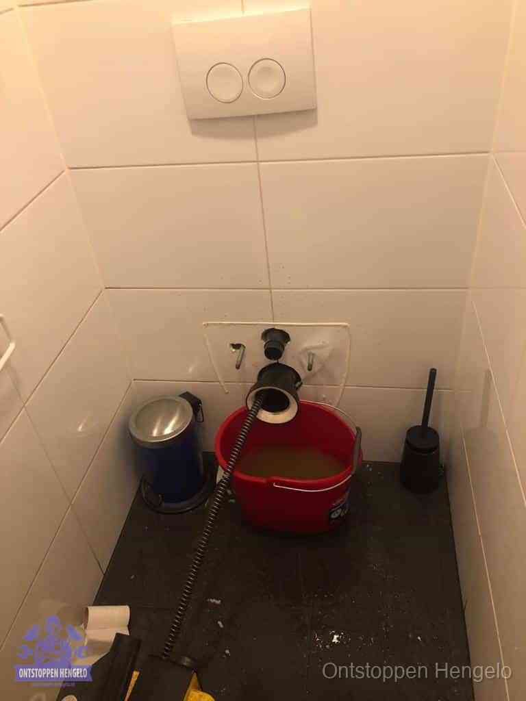 Een ontstopte wand-wc met verwijderde toiletpot waar een ontstoppingsveer in de wanduitgang verdwijnt, het achterhalen waar de verstopping zit in Hengelo