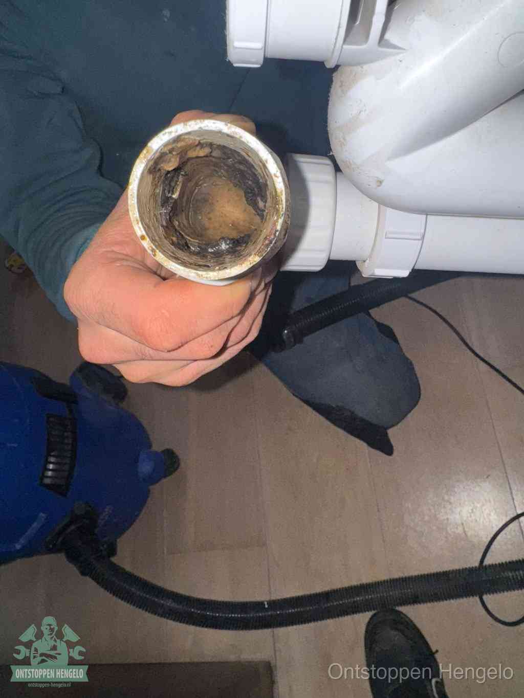 Een hand houdt een losgekoppeld PVC-afvoerbuisstuk vast dat van binnen volledig is dichtgeslibd met vet, vuil en aanslag, de meest voorkomende oorzaak van een verstopte afvoer in Hengelo