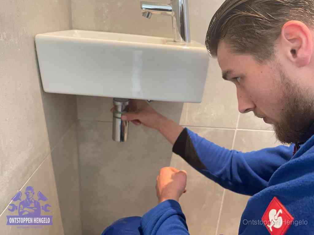 Een monteur draait met de hand de zwanenhalssifon onder een fonteinwasbak in de toiletruimte los om de bron van rioollucht in Hengelo te vinden
