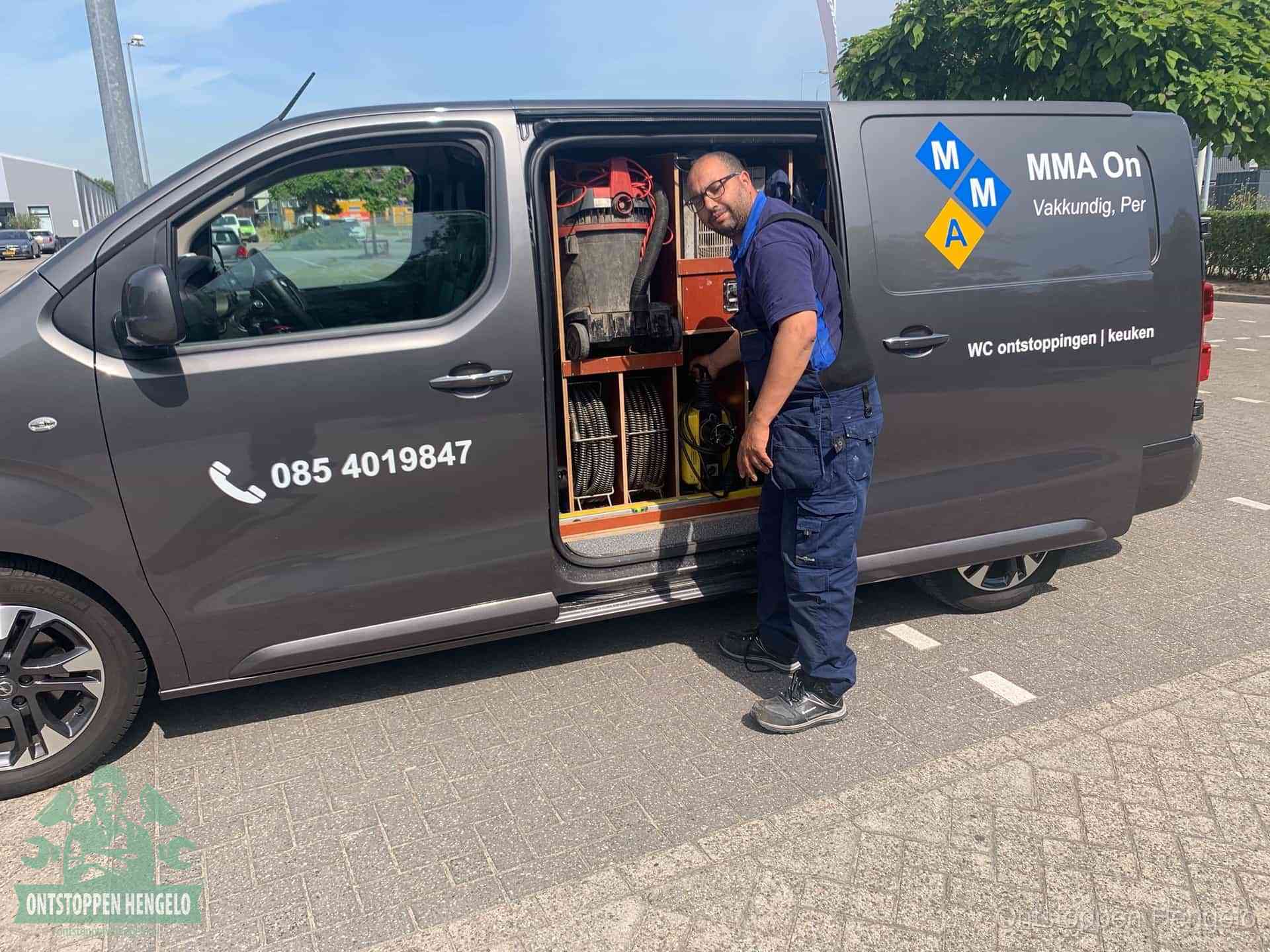Een monteur bij de open schuifdeur van zijn bedrijfsbus op een parkeerplaats, klaar om de oorzaak van terugkerende afvoerverstoppingen aan te pakken in Hengelo