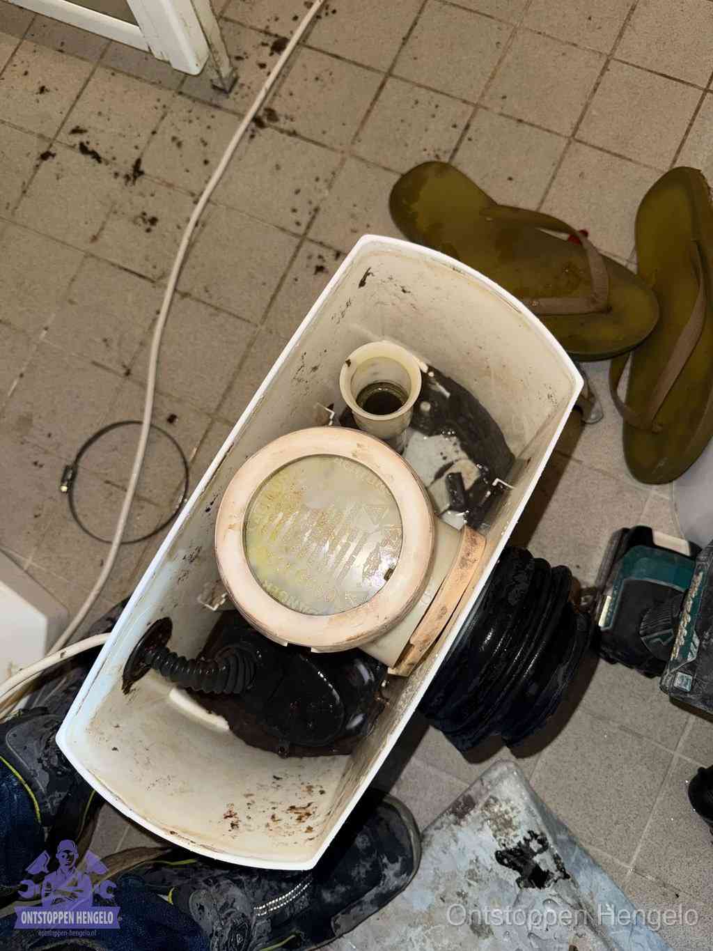 Een gedemonteerde toiletpot op de vloer met ontstoppingsveer en gereedschap ernaast, het verwijderen van een vastzittend voorwerp uit de wc in Hengelo