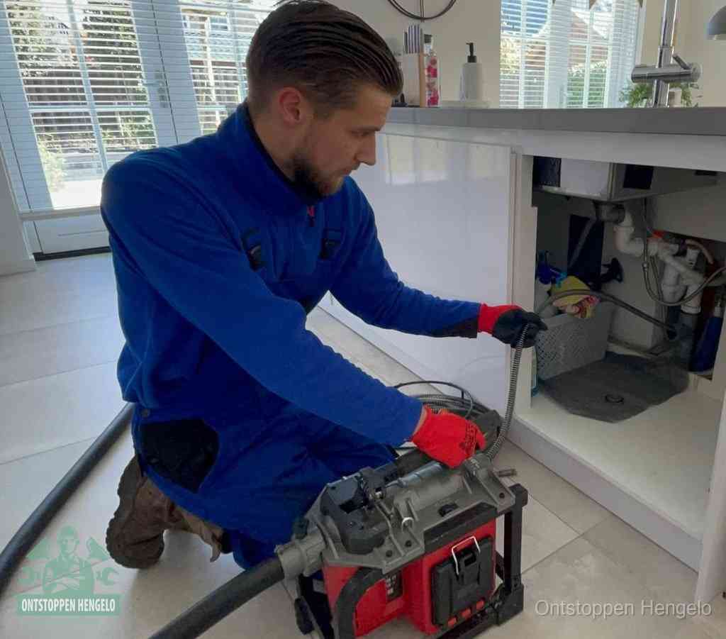 Een monteur met rode handschoenen knielt in een keuken en voert een elektrische ontstoppingsveer in tijdens het professioneel afvoer ontstoppen in Hengelo